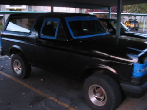 1991 Ford Bronco - A black SUV is parked under a covered area, with blue tape applied to its windows and front end.