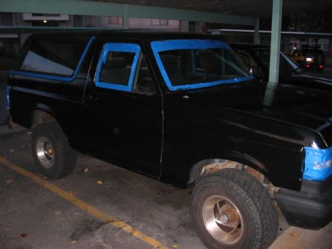 1991 Ford Bronco - A black SUV, likely a Ford Bronco from the early 1990s, is parked in a lot with blue tape applied to its windows and lights.