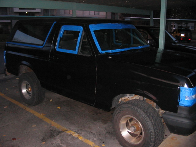 Black SUV, possibly a Ford Bronco, with blue tape around windows and headlights. Visible rust on wheel wells. Parked in a lot