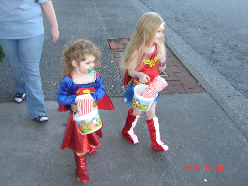 Two kids in superhero costumes, one as Superman and one as Wonder Woman, walk down a sidewalk holding candy buckets