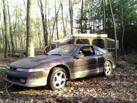 1976 GMC C/K Pickup - A dusty coupe sits in a wooded area next to a pop-up camper, suggesting a campsite or abandoned location.