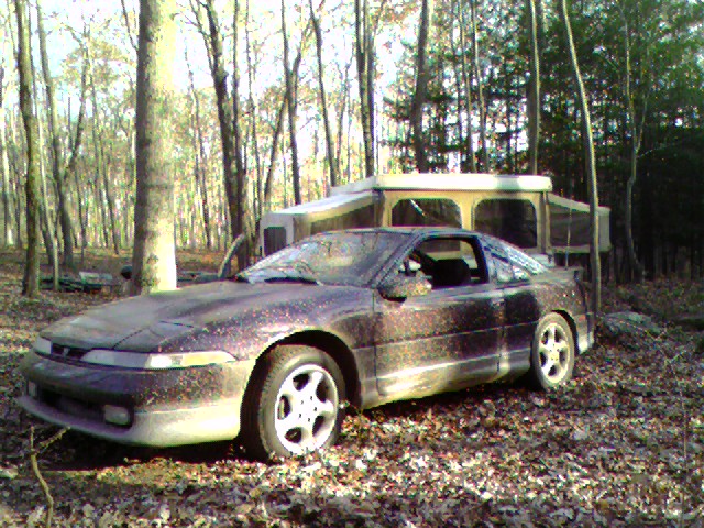 Dark coupe with a pop-up camper in a wooded area. The car is covered in dust and has aftermarket wheels