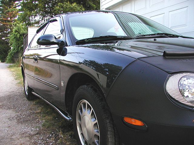 Dark Ford Taurus sedan with black front-end bra on gravel driveway next to white garage