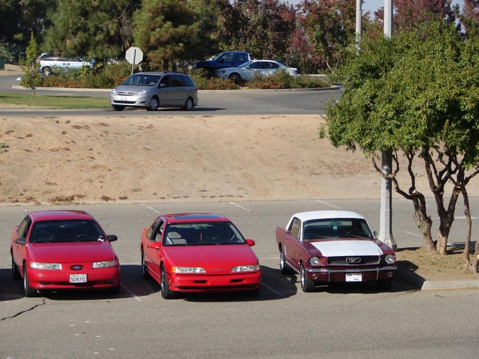 Three red cars parked in a lot, one is a 1965 Ford Mustang with a white top. A silver van is driving in the background