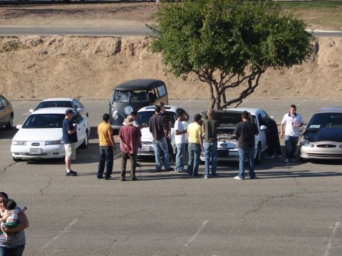 1992 Ford Taurus - Several vehicles and a group of people are gathered in a parking lot, with some individuals inspecting the engines of the cars.