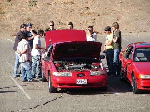 1992 Ford Taurus - Group of people examining the engine of a red 1992 Ford Taurus at an outdoor gathering.