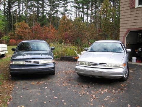 1991 Ford LTD Crown Victoria - Two Ford Crown Victorias are parked outside a garage on a fall day, with fallen leaves on the ground.