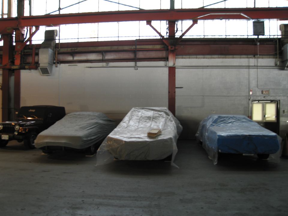 Three cars covered in plastic sheeting parked indoors, with a black Jeep visible on the left