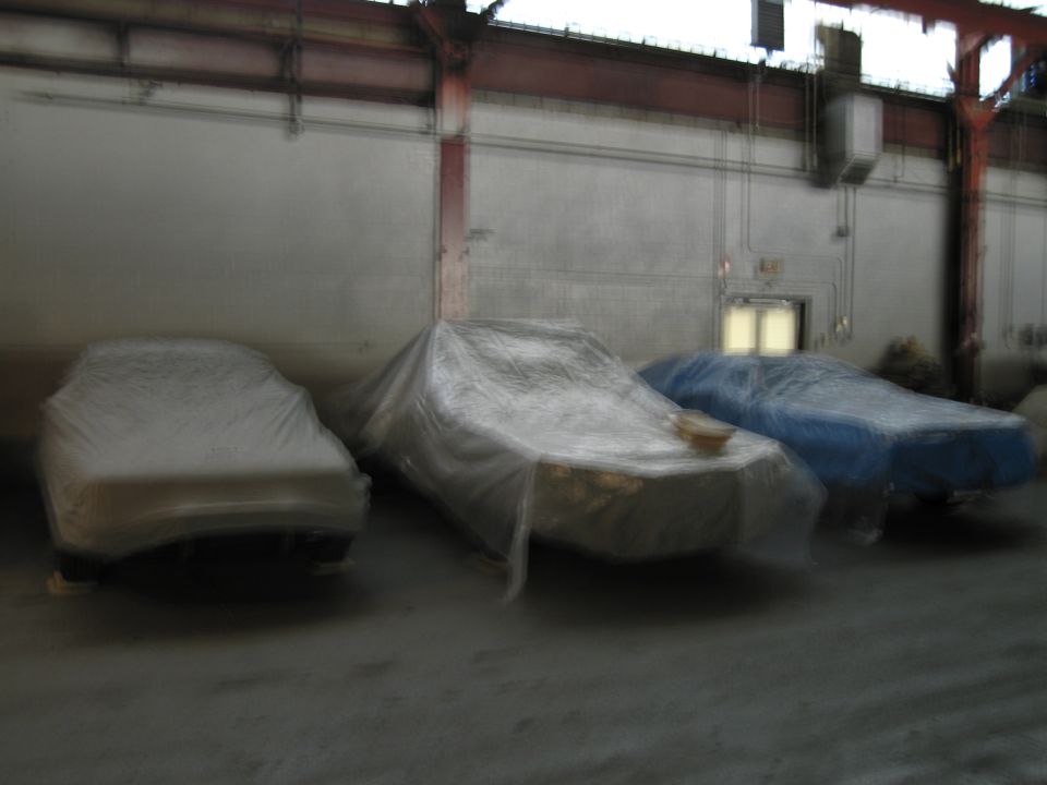 Three cars covered in tarps sit in a dimly lit warehouse. One is white, one is tan, and one is blue