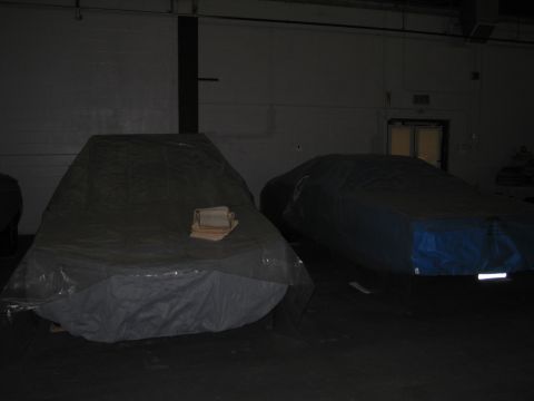 1991 Ford LTD Crown Victoria - Two vehicles are covered with tarps in a dimly lit indoor space, with a stack of papers on the tarp of the vehicle on the left.