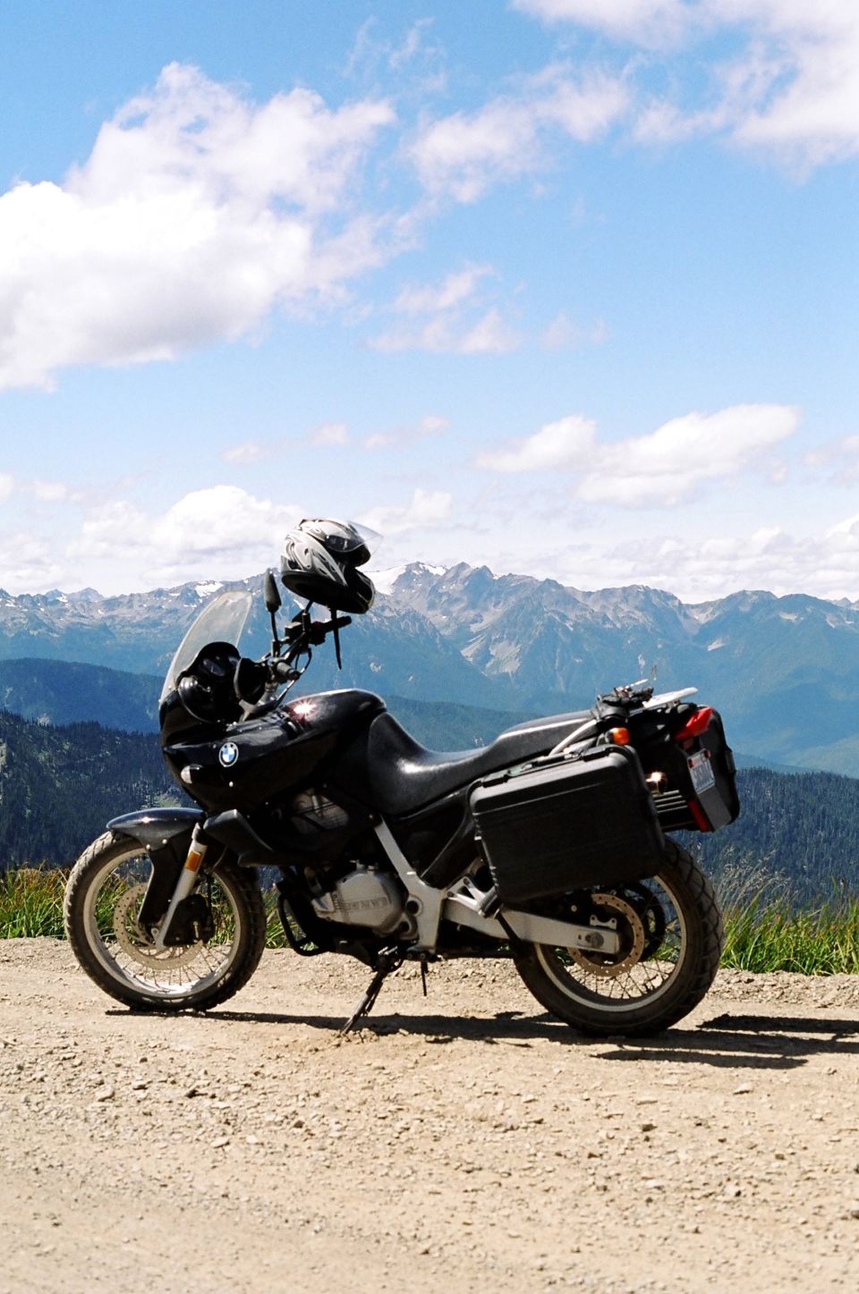 Black motorcycle with saddlebags parked on a dirt road, mountains in background. Helmet on handlebars