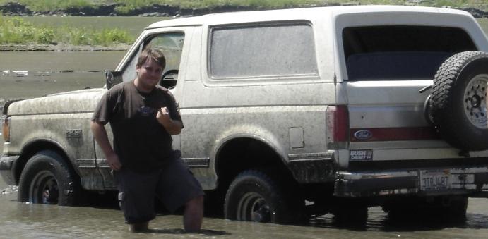 Muddy white Ford Bronco parked in shallow water with a man standing knee-deep beside it