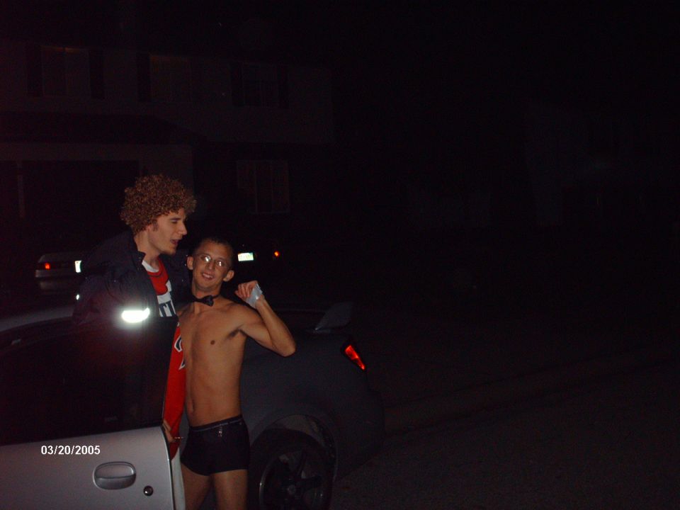 Two guys, one in a wig, pose next to a car at night. Date stamp reads 03/20/2005