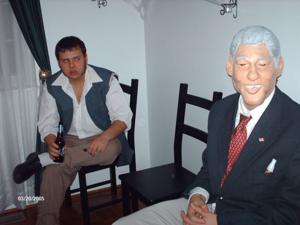 Guy in vest and colonial shirt holds beer, sitting next to a Bill Clinton mask on a person in a suit