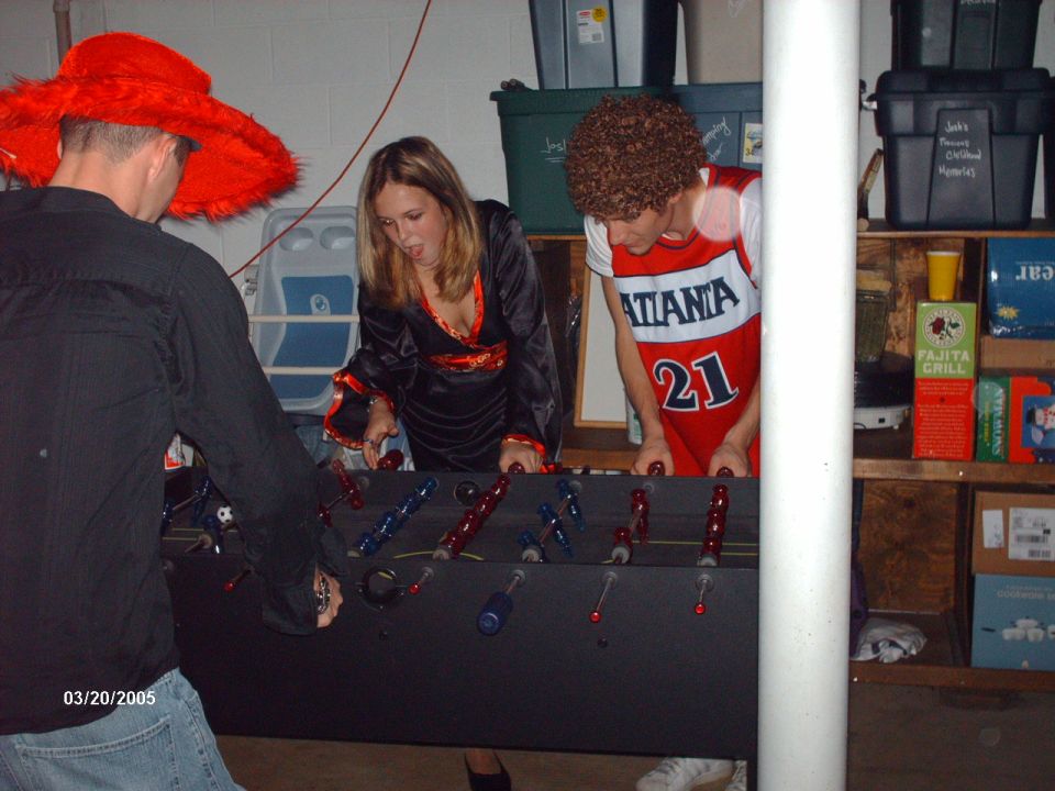 Three people in costumes play foosball in a basement. One wears a large red hat, another a black robe, and the third a basketball jersey