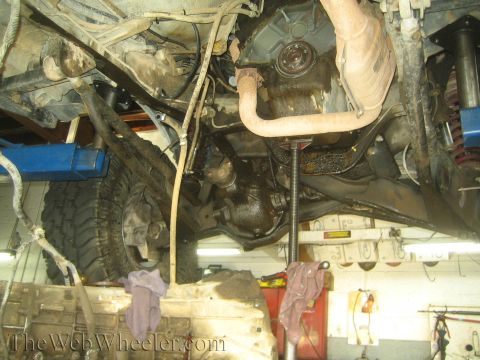 1990 Ford Bronco - Underbody view of a vehicle on a shop lift showing the engine pan, oil filter, exhaust pipe and a jack supporting the drivetrain.