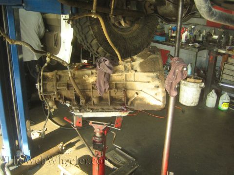 1990 Ford Bronco - automatic transmission lowered onto a red transmission jack beneath a raised vehicle in a repair shop.