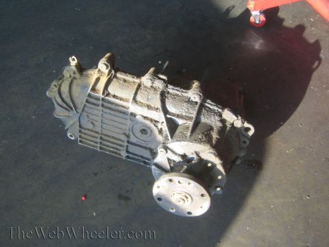 1990 Ford Bronco - dirty cast-aluminum transfer case removed from a vehicle and sitting on a dark shop floor near a jack.
