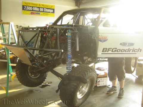 1990 Ford Bronco - white tube-chassis off-road race truck raised on a shop lift with blue coilover shocks and BFGoodrich lettering, a person standing alongsid…