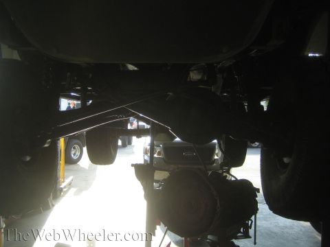 1990 Ford Bronco - Backlit underbody view of a vehicle on a lift showing the rear axle, driveshaft and suspension silhouetted against shop lights.