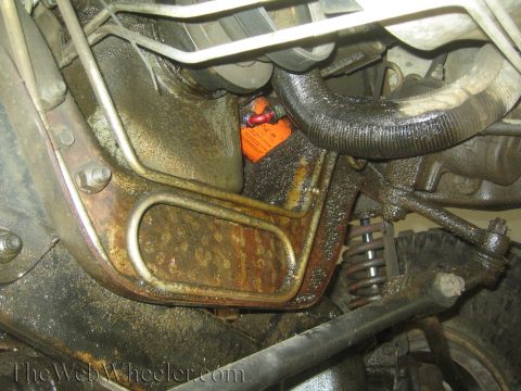 1990 Ford Bronco - Close-up of a rusty vehicle frame and bracket with an orange tag, heater hose and coil spring visible underneath.
