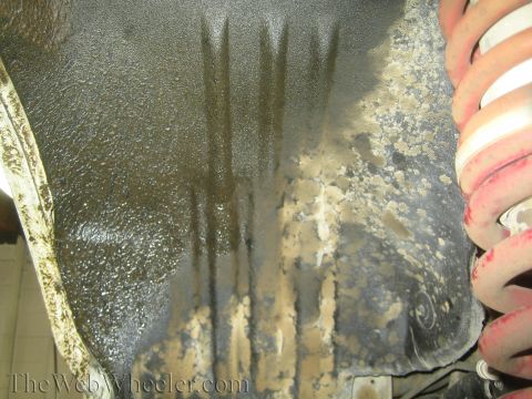 1990 Ford Bronco - Underside view of a greasy, dirt-caked black metal component with a red-tipped coil spring visible at right.