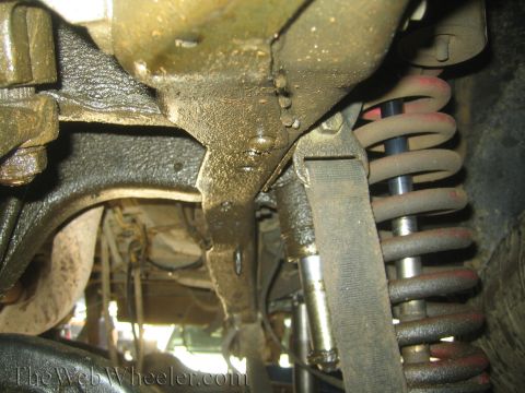1990 Ford Bronco - Underside of a vehicle showing a dirty front axle assembly, a red-painted coil spring, and a steering linkage.