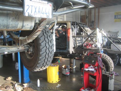 1990 Ford Bronco - tube-chassis race truck sits on a shop lift, its bodywork stripped, with a knobby off-road tire and visible license plate.