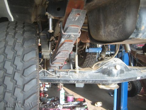 1990 Ford Bronco - Underside of a vehicle on a lift showing the frame rail, leaf spring shackle mount, fuel tank, and an off-road tire.