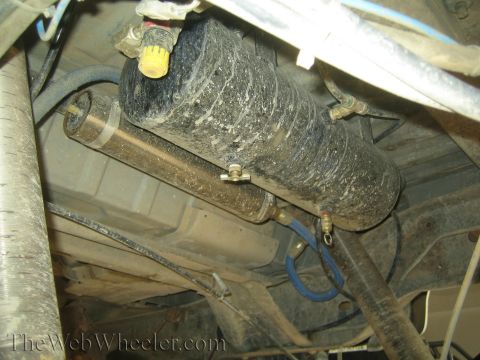 1990 Ford Bronco - Underside closeup of a heavily soiled cylindrical canister, likely a muffler, surrounded by hoses and wiring.