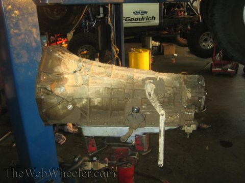 1990 Ford Bronco - removed automatic transmission case rests on a jack in a shop, with a BFGoodrich banner and vehicles in the background.