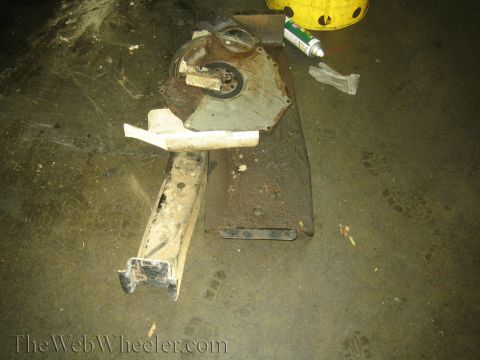 1990 Ford Bronco - removed bellhousing dust cover plate and a crossmember rest on a dirty shop floor beside a tube of sealant.