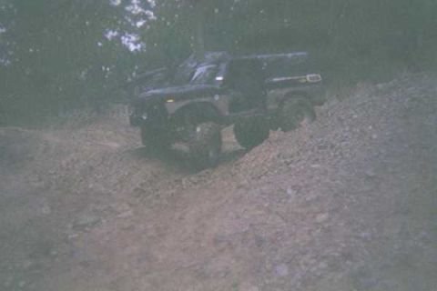 1978 Ford Bronco - A dark-colored off-road vehicle navigates a steep dirt incline surrounded by trees.