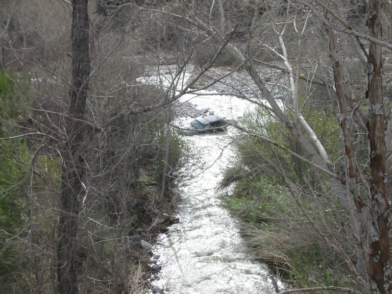 Distant view of a dark SUV submerged mid-stream in a swollen whitewater creek seen through bare trees