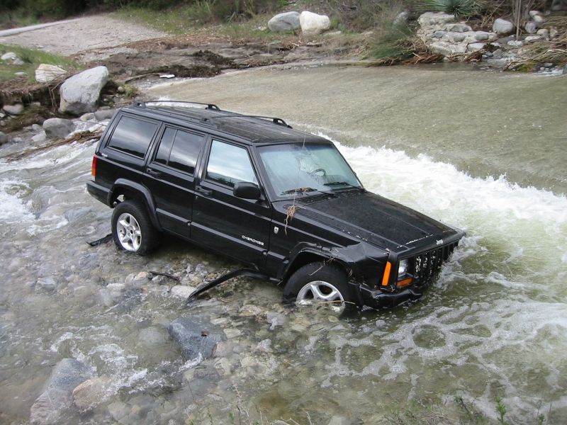 Passenger side of a black Jeep Cherokee XJ sitting in a rocky river with a damaged front fender