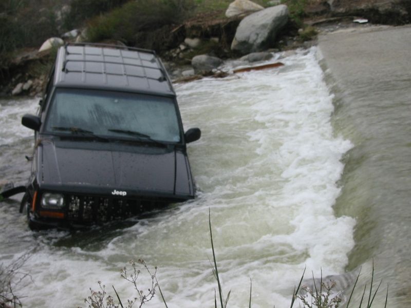 Dark gray Jeep Cherokee XJ stuck half-submerged in whitewater at a concrete spillway