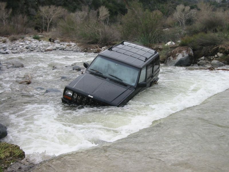 Black Jeep Cherokee XJ stranded nose-down in a fast silty river between boulder banks