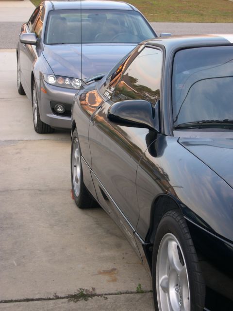 1997 Ford Probe - Two cars parked nose-to-nose on a concrete driveway at dusk: a glossy black coupe in the foreground shot from a low front-quarter angle ove…