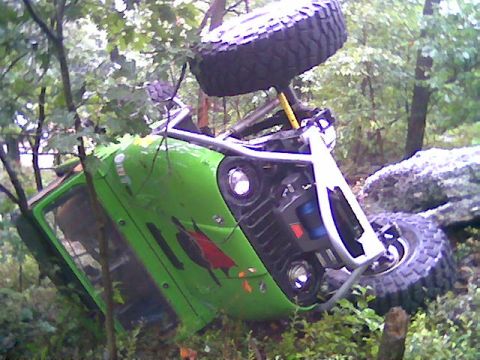 1976 GMC C/K Pickup - An off-road vehicle has overturned in a forest, resting on its side amongst trees and rocks.