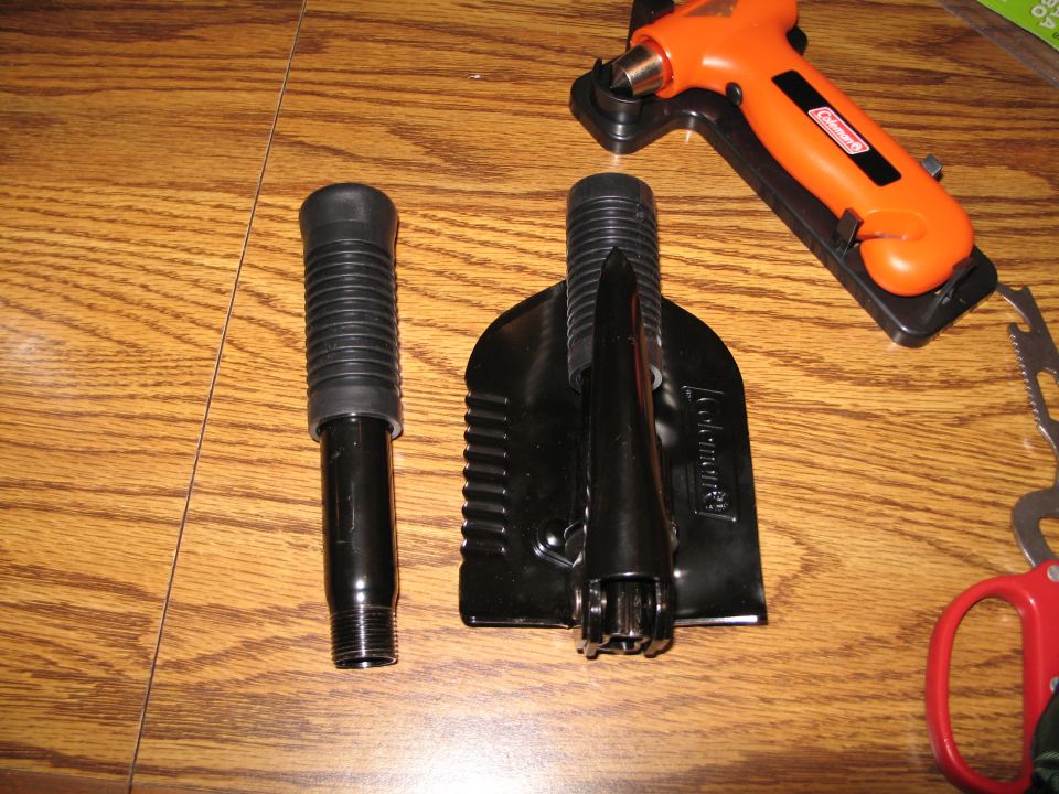 Camping shovel with handle, Coleman multi-tool with hammer and seatbelt cutter, and red handled scissors on a wooden surface