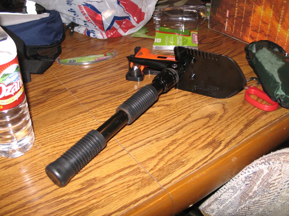 Black folding shovel with orange accents on a wooden table next to a water bottle and bags