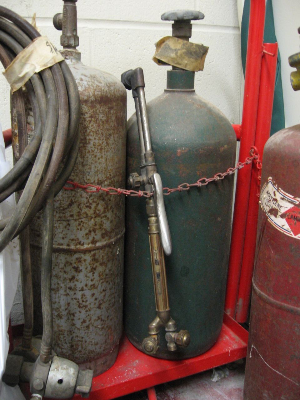 Gas cylinders and a welding torch on a red cart. One cylinder is green, another is rusty, and a third is red with a label