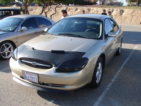 1992 Ford Taurus - A gold sedan with a black car bra and aftermarket headlights is parked in a lot next to another car.