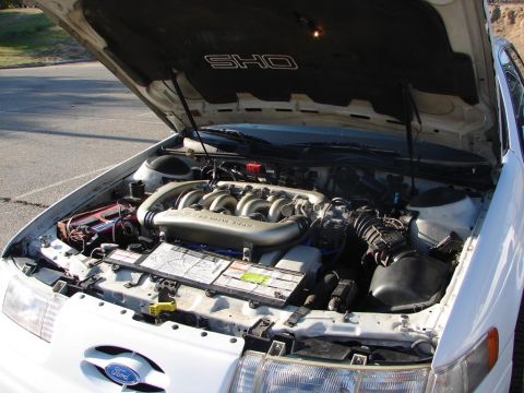 1992 Ford Taurus - The engine bay of a white 1992 Ford Taurus SHO is visible with the hood open, revealing the V6 engine and 'SHO' badging.