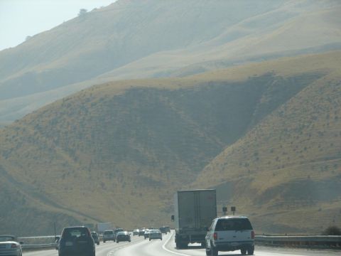 1992 Ford Taurus - Traffic moves along a highway flanked by dry, rolling hills under a hazy sky.
