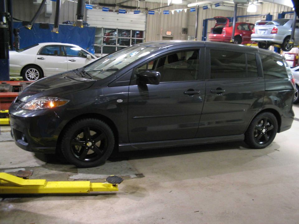 Dark gray Mazda5 minivan on black alloy wheels parked inside a repair shop