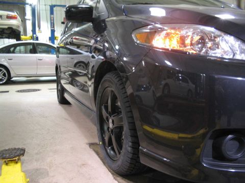 2006 Mazda Mazda 5 - A dark 2006 Mazda Mazda 5 is shown in a service bay, with another vehicle on a lift in the background.