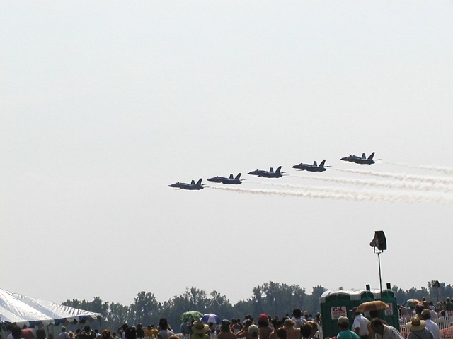 Five fighter jets fly in formation, leaving contrails against a hazy sky. A crowd watches from below