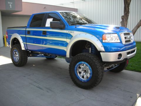 1998 Ford Crown Victoria - lifted blue Ford F-150 crew-cab pickup on oversized mud tires and blue beadlock wheels sits on a driveway by a building.