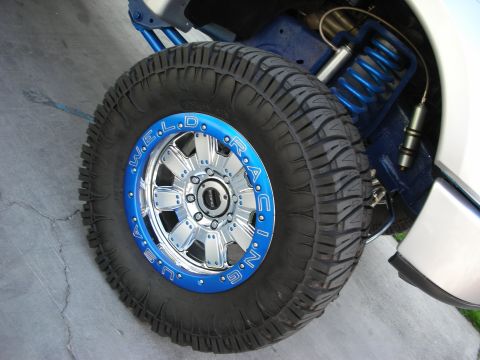 1998 Ford Crown Victoria - close-up shows a Weld Racing wheel with a blue beadlock ring and chunky mud tire mounted on a lifted truck with blue suspension.